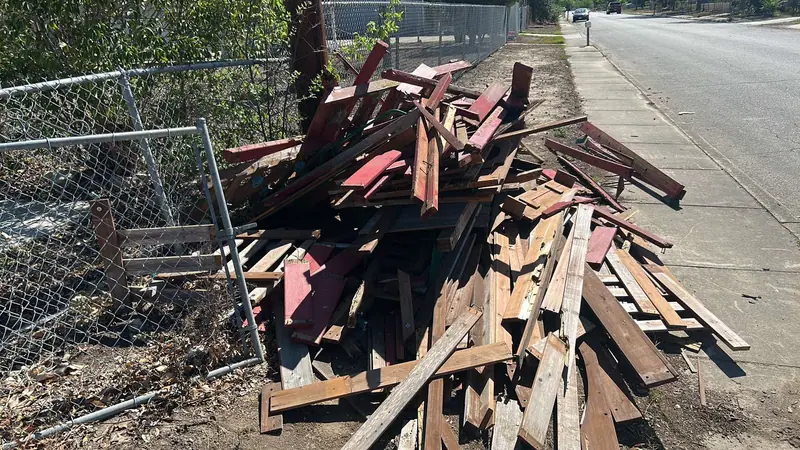 Curbside pile of demolished red stained deck lumber and framing beside a chain link fence