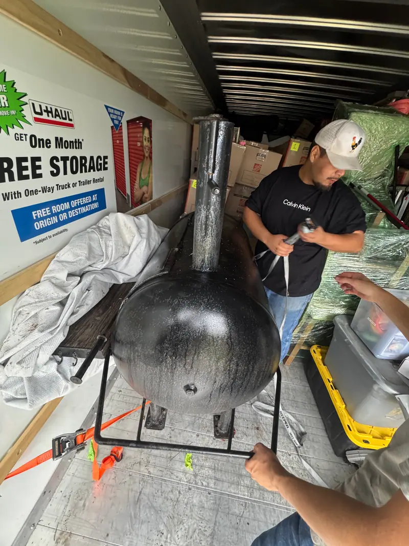 Two crew members loading a large steel offset BBQ smoker into a box truck