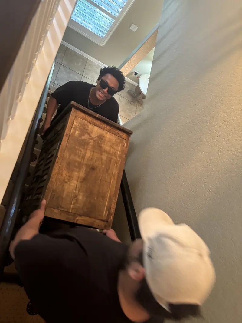 Two Elite Demo and Junk Removal crew members carrying a heavy wooden dresser down a narrow staircase in San Marcos