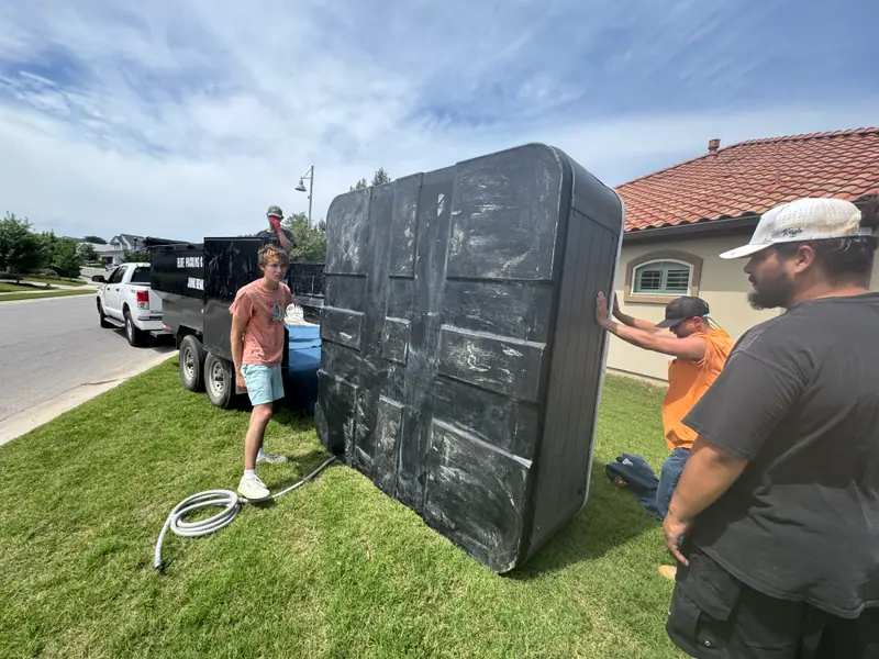 Elite Demo and Junk Removal crew standing a hot tub shell upright beside the company dump trailer in San Marcos