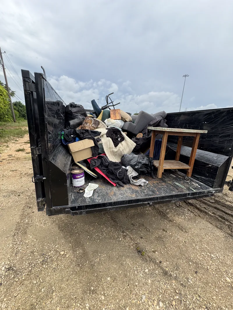 Dump trailer loaded with household junk including a chair, bagged waste and a wooden side table