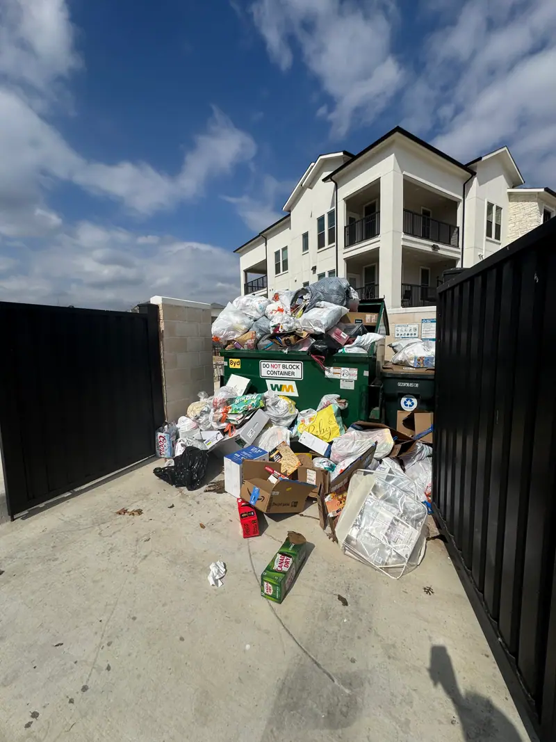 Overflowing apartment dumpster enclosure in San Marcos with bulk items spilling onto the pad